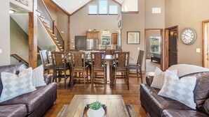Uma sala de estar espaçosa com pé direito alto, uma área de jantar com mobiliário de madeira e uma cozinha ao fundo.
