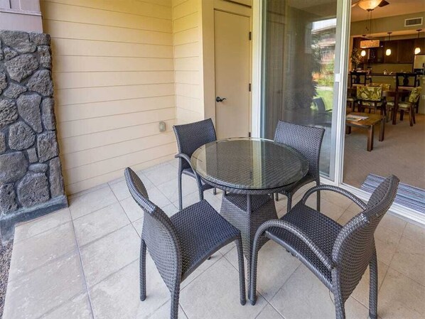 Een terras met een ronde tafel en vier rieten stoelen, een stenen muur en een glazen deur naar een eethoek binnen.