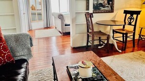 Un salon doté d’un plancher en bois, d’un canapé, d’une table basse avec un magazine et un objet décoratif, d’un tableau au mur et d’un coin repas avec des chaises et une table.