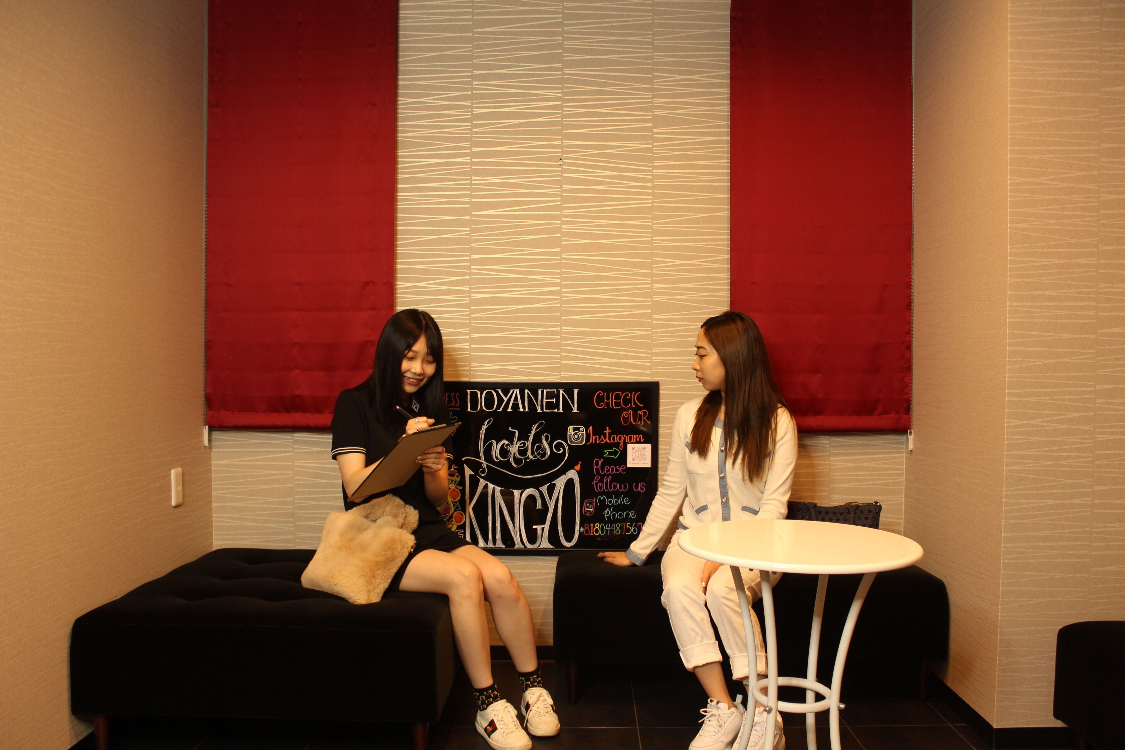 Two individuals seated in a room with a red accent wall, a black couch, and a small round table. A sign with text and social media information is visible.