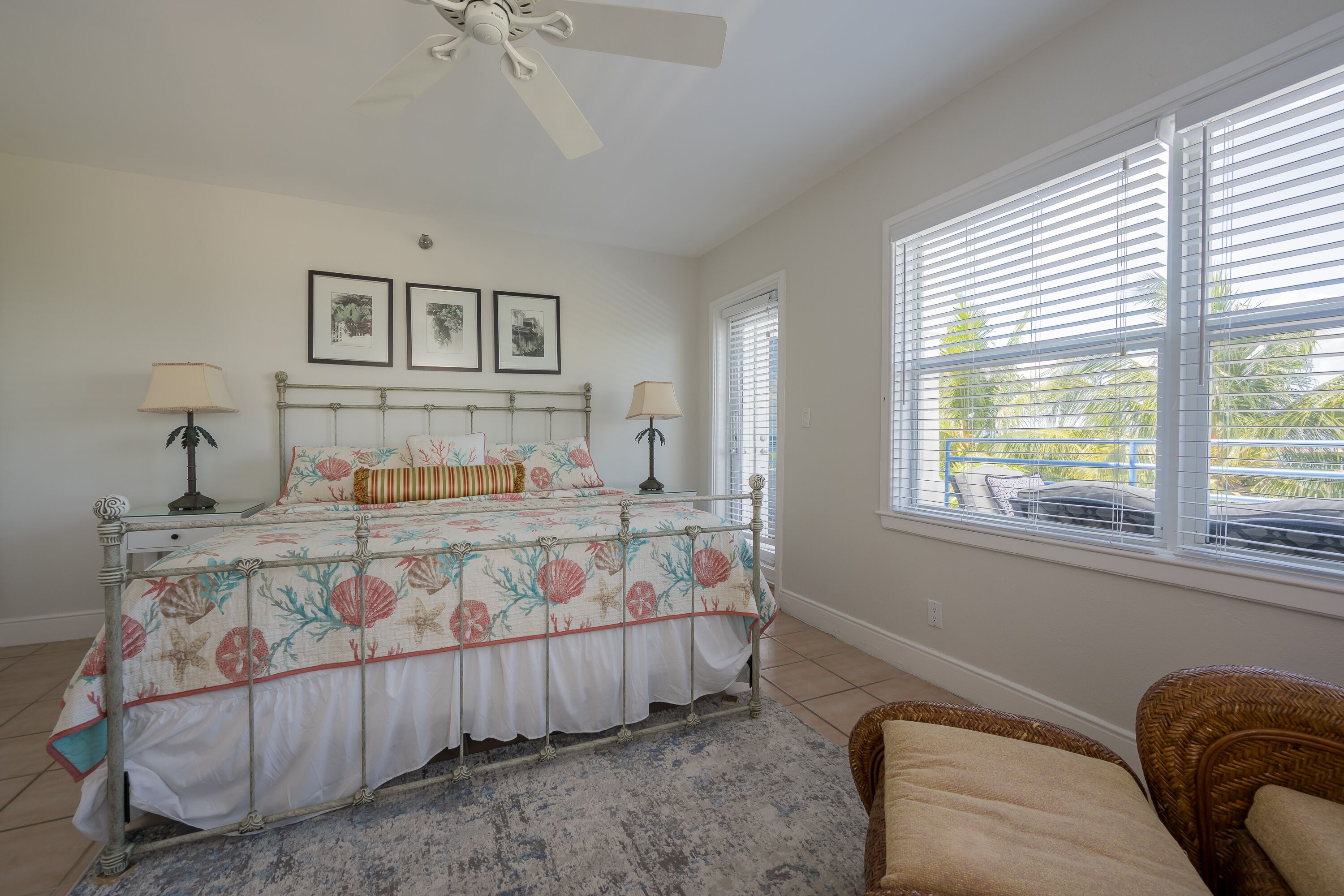 A bedroom with a bed, two bedside lamps, a ceiling fan, a window with blinds, and a view of the outdoors.