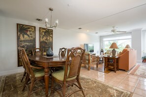A dining area with a round table, chairs, and a chandelier. There is a sofa and a television in the background.