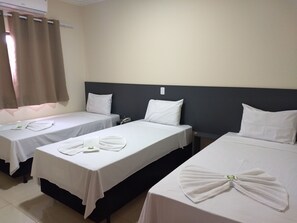 Habitación de hotel con tres camas individuales, cada una con sábanas y almohadas blancas. Hay una mesita de noche con teléfono y una lámpara pequeña. La habitación tiene una decoración en tonos neutros, con paredes beige y un cabecero oscuro.