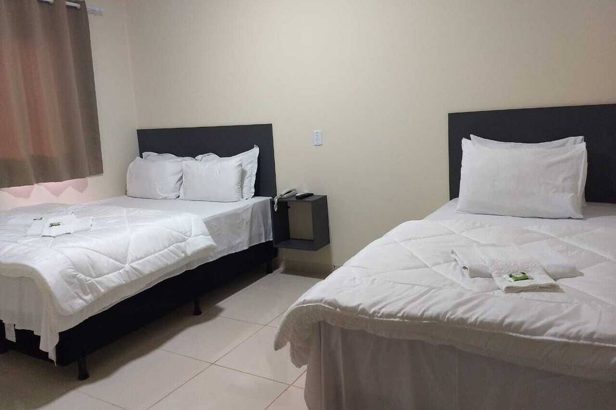 A hotel room with two single beds, each with white bedding and headboards. There is a small bedside table with a lamp and a phone on it. The room has a plain wall and a curtain on the window.