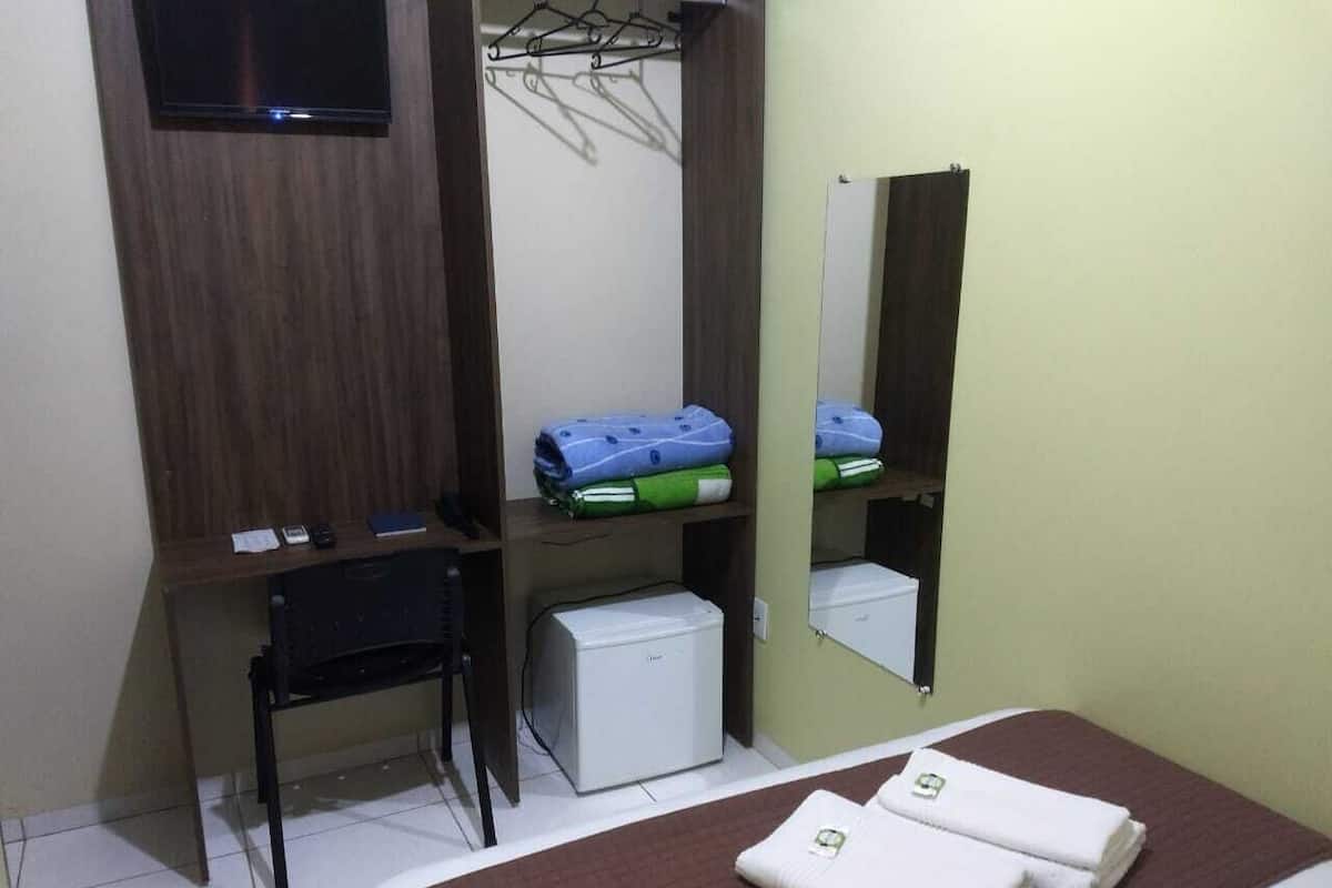 A hotel room with a bed, a desk, a chair, a small refrigerator, and a mirror.