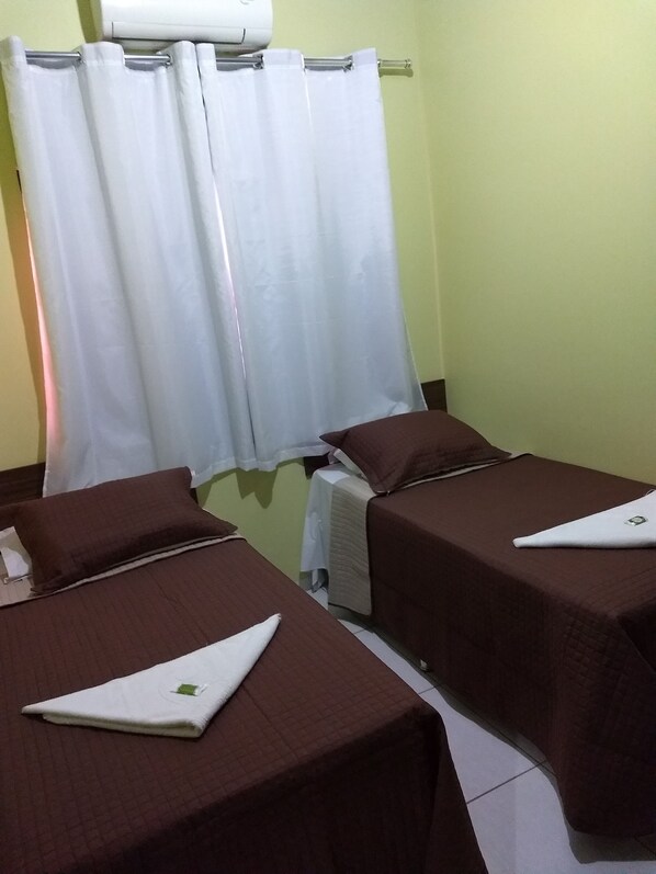 Una habitación con dos camas, cada una con una colcha marrón y una almohada blanca. Hay una cortina blanca en la ventana, y un equipo de aire acondicionado blanco encima.
