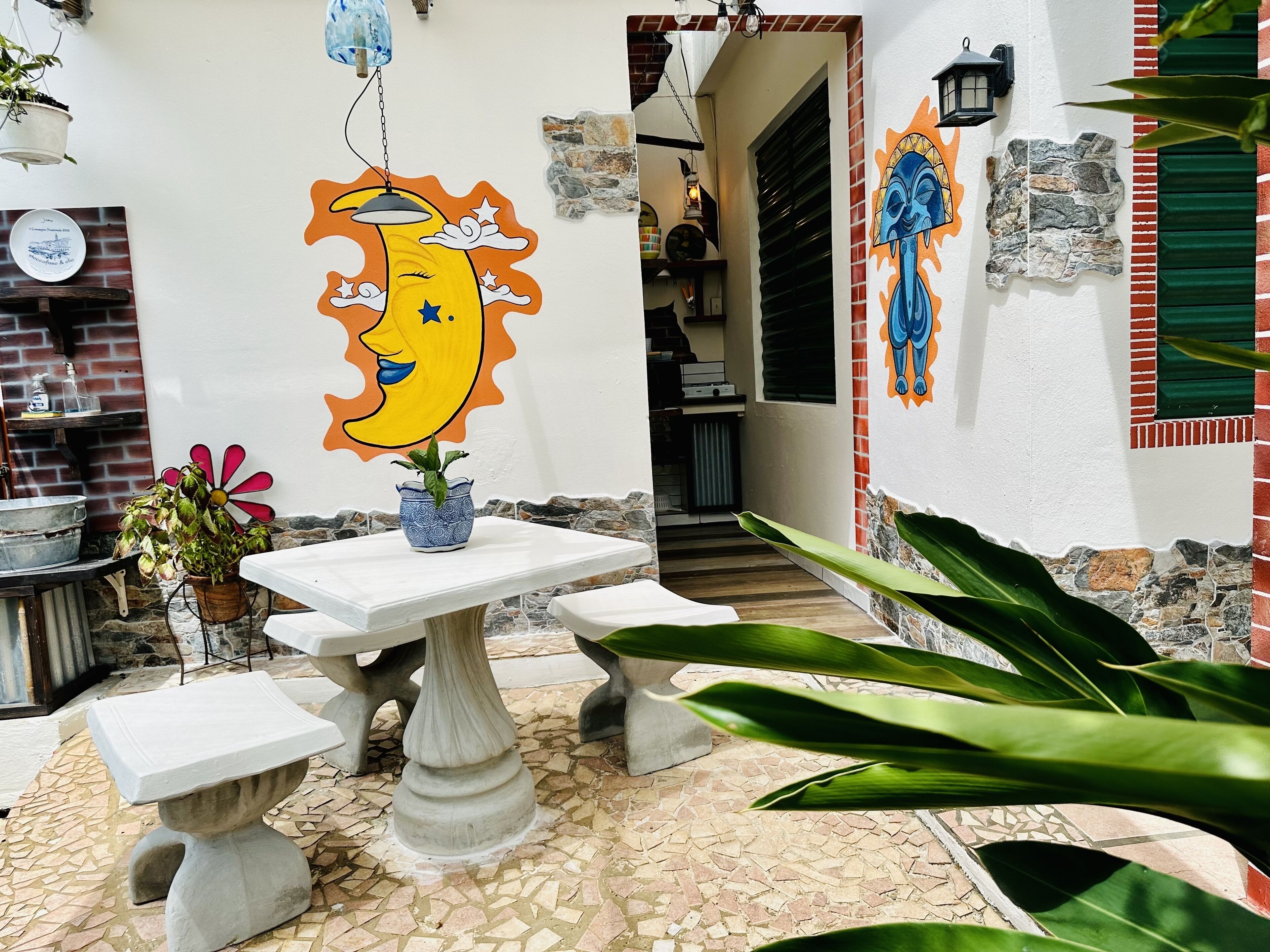 A courtyard with a white table and benches, a potted plant, and a mural of a smiling moon on the wall.
