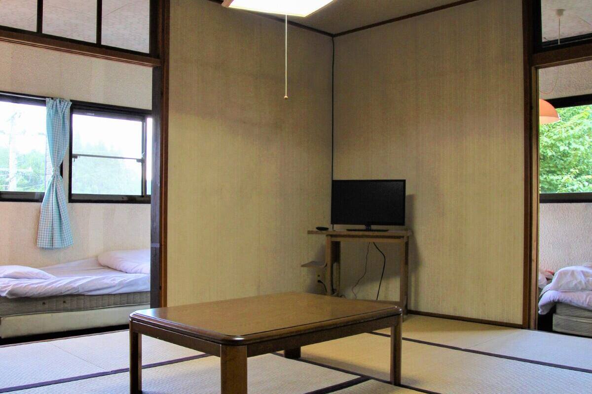 A traditional Japanese room with tatami flooring, a low wooden table, a television on a stand, and a window with a curtain.