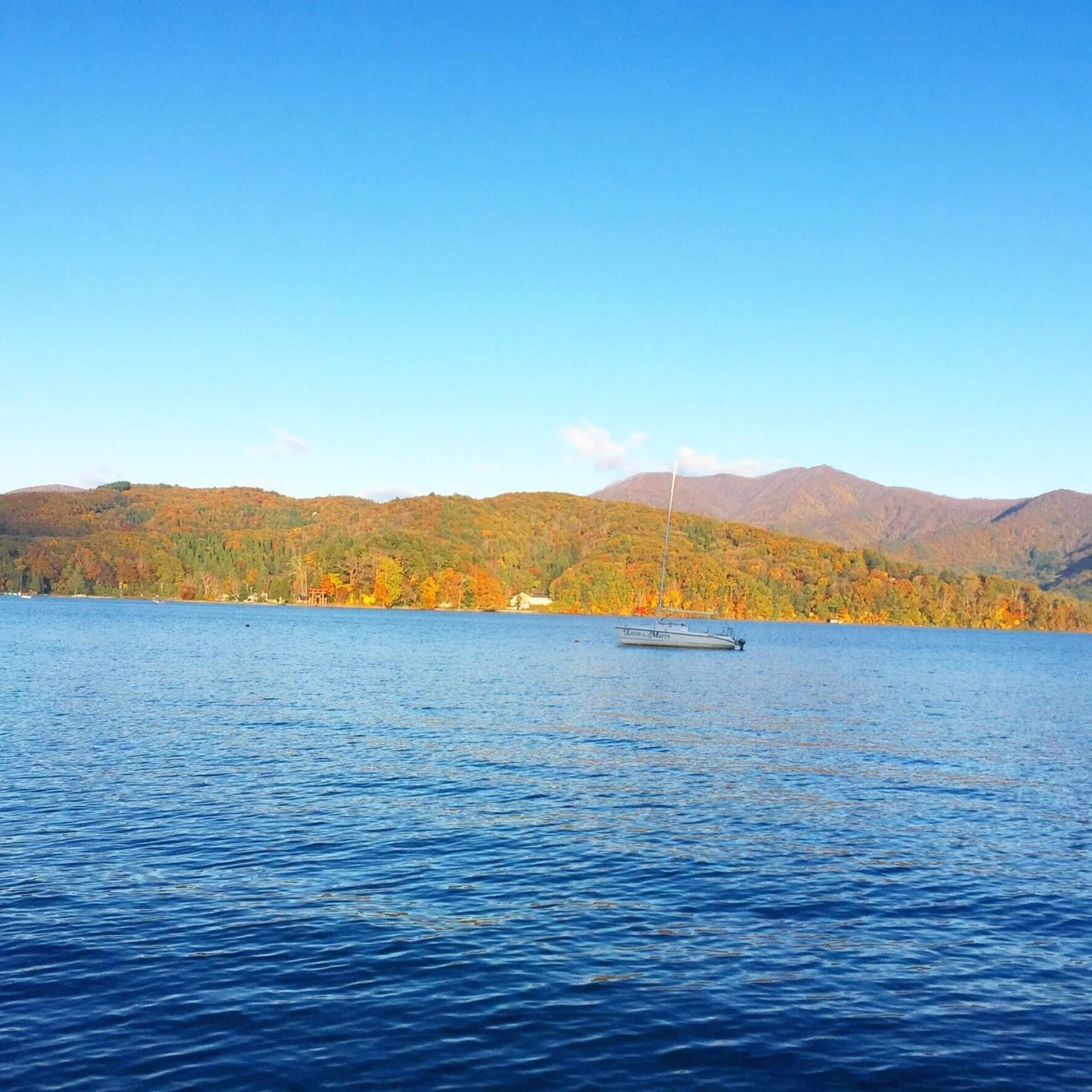 Un lago tranquilo con un velero en el centro, rodeado de árboles de colores otoñales y montañas al fondo.