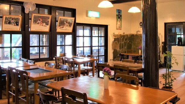Interior de restaurante con mesas y sillas de madera, ventanas amplias y lámparas colgantes.