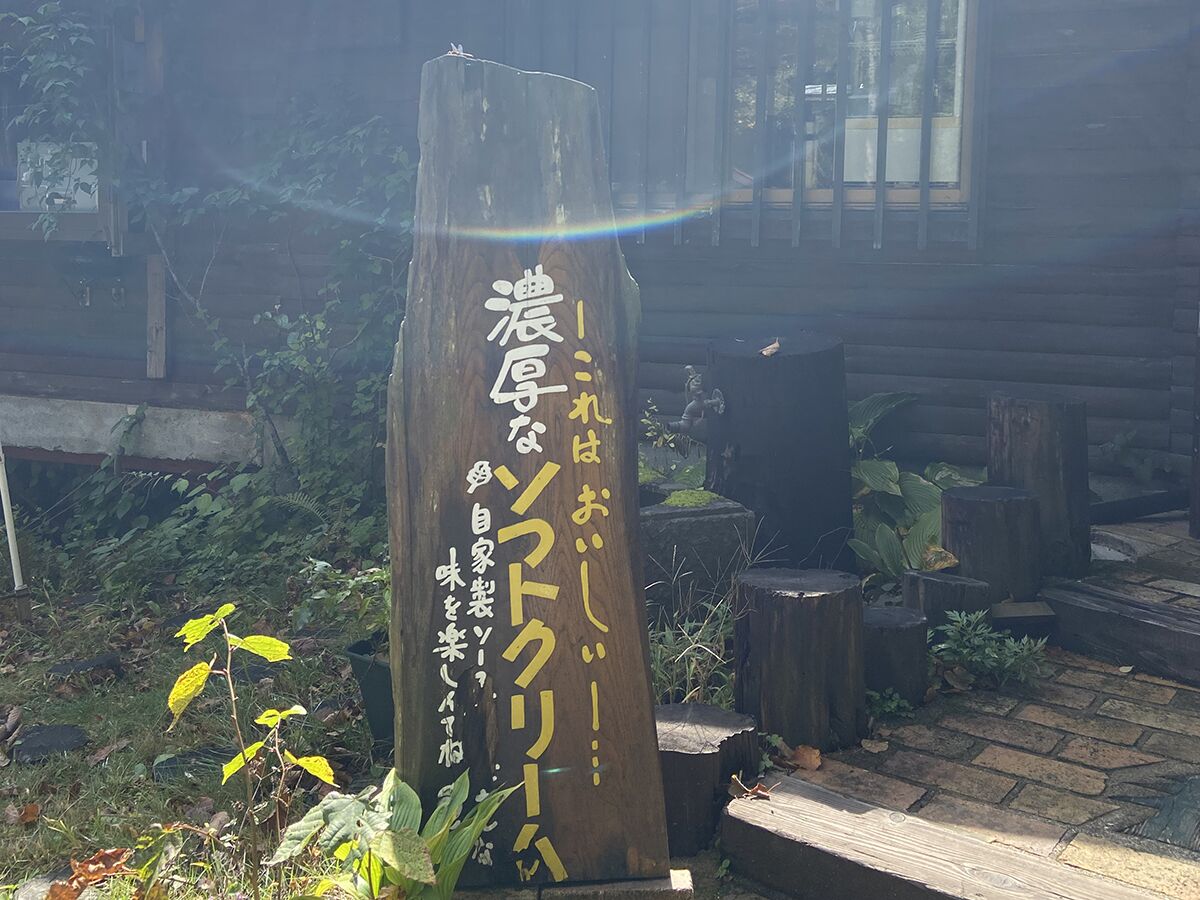 Un letrero de madera con caracteres japoneses, rodeado de follaje y una estructura de madera al fondo.