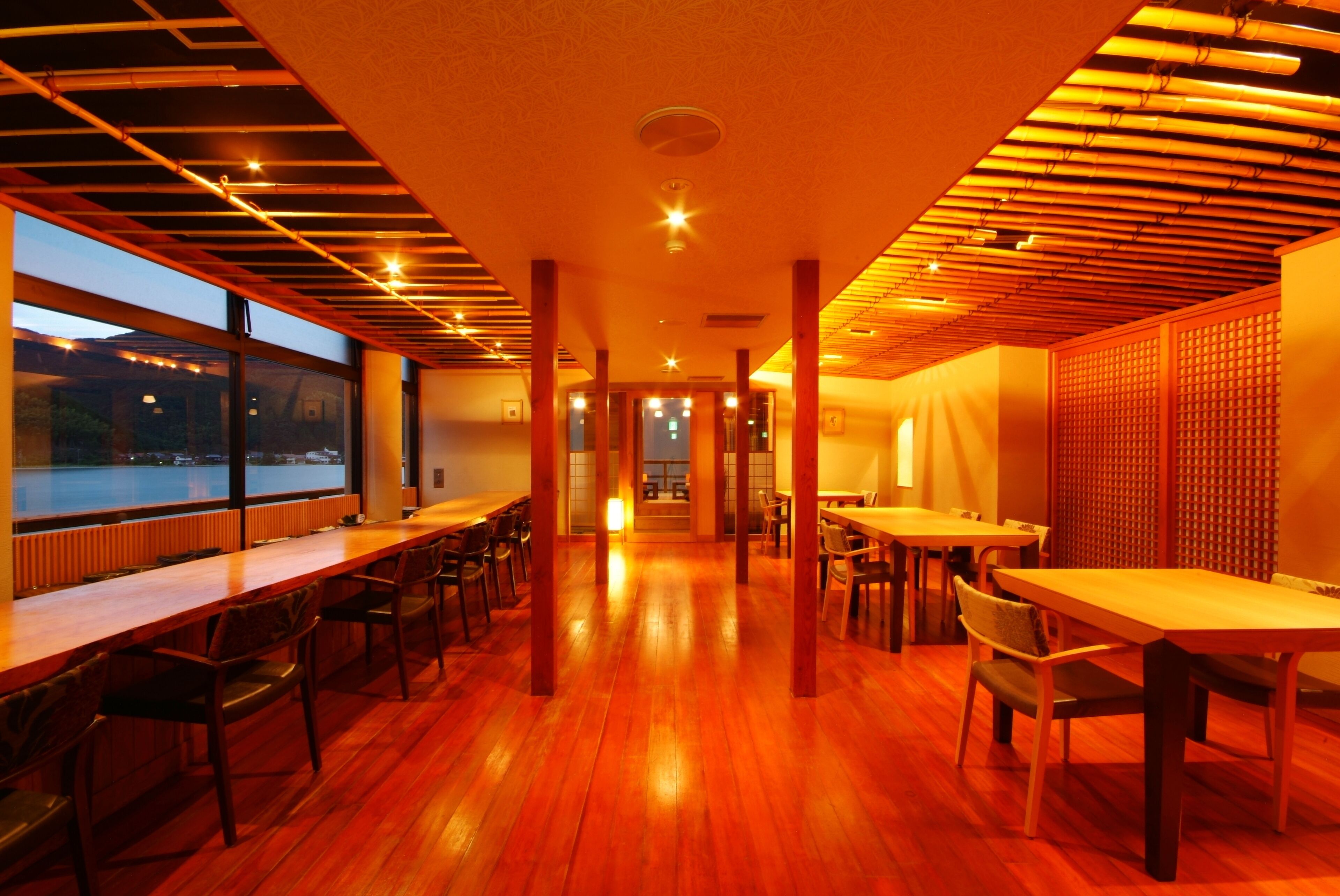A restaurant with wooden floors and tables, large windows, and a view of the water outside.