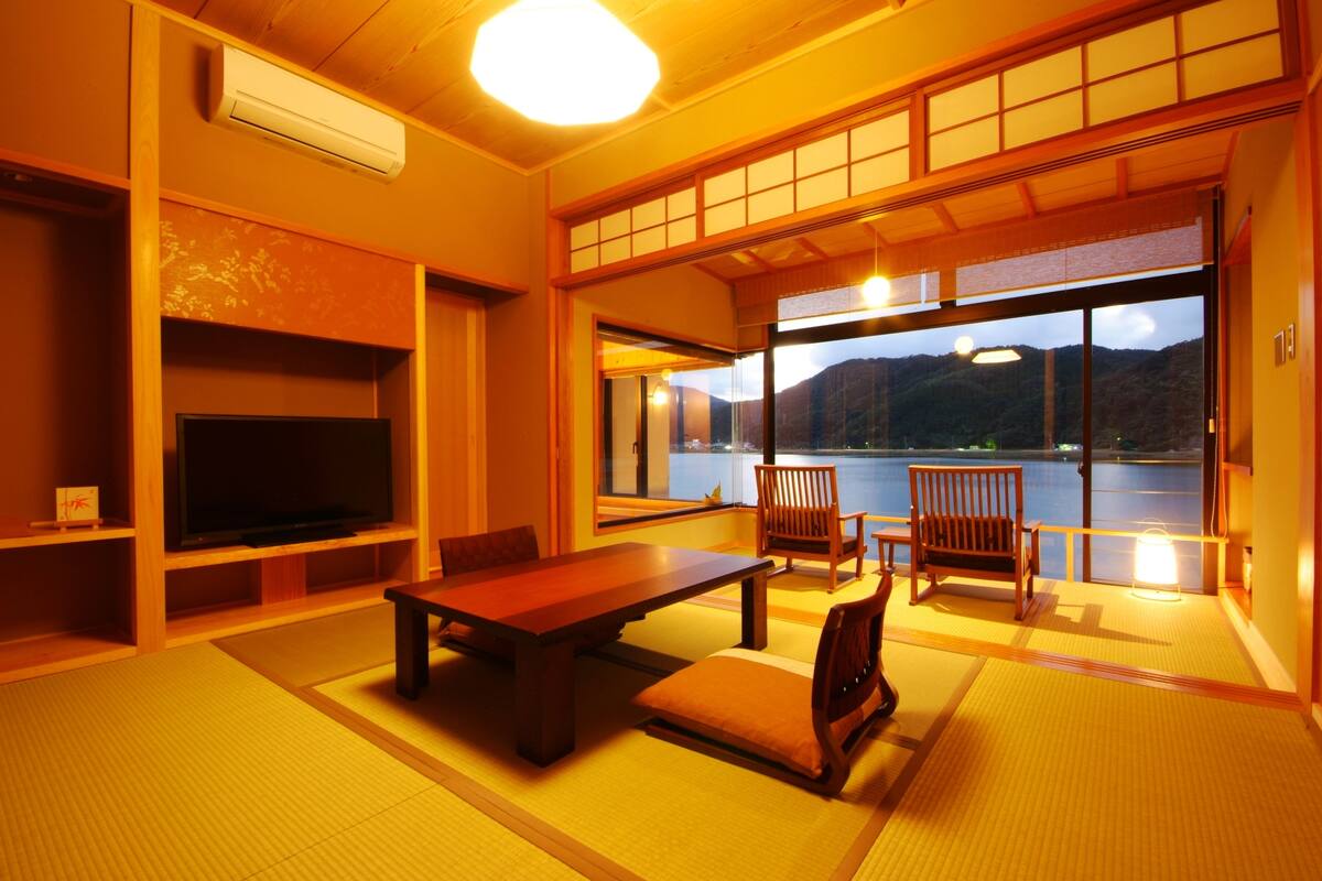 A traditional Japanese room with tatami flooring, a low table, chairs, a television, and a view of a lake and mountains.