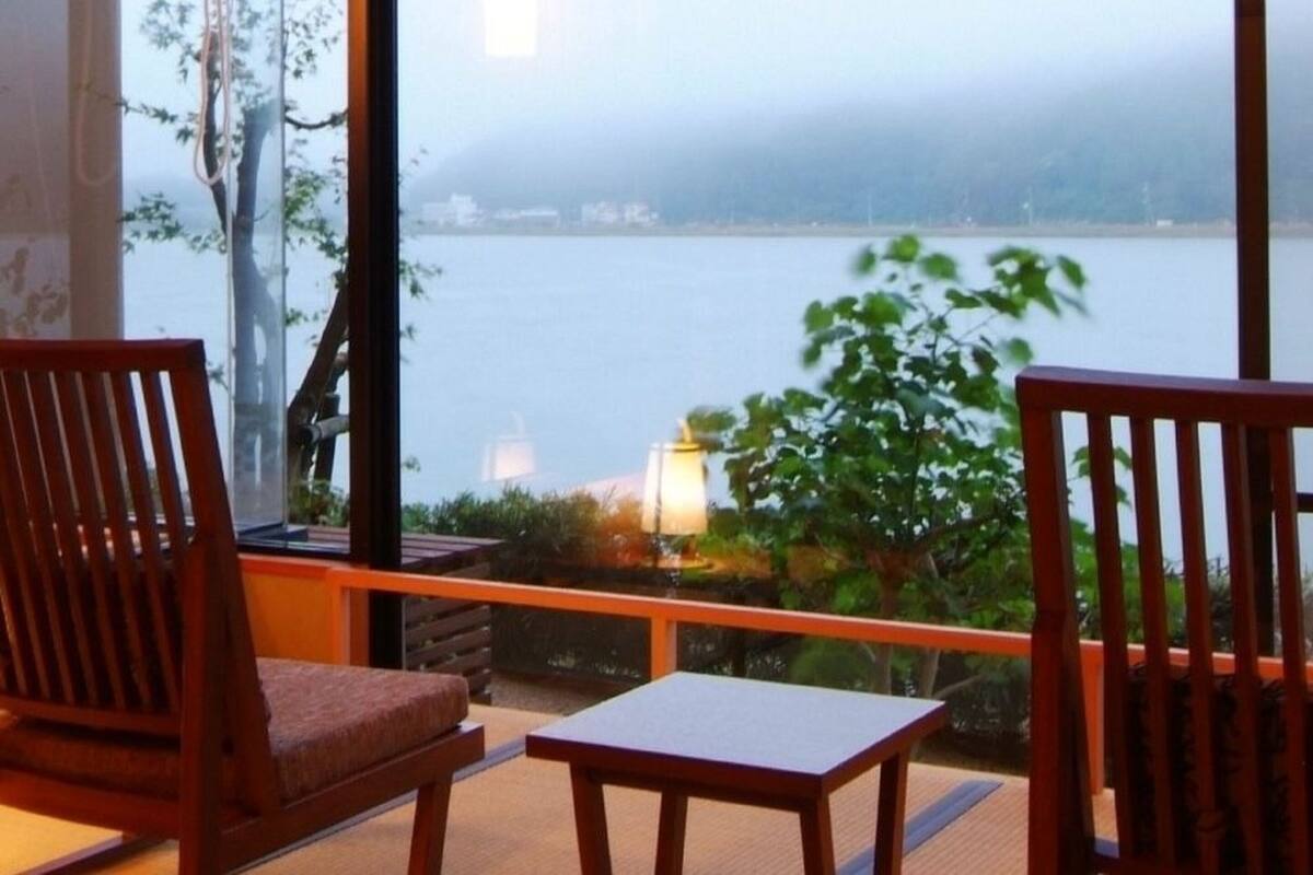 A balcony with wooden chairs and a table, overlooking a body of water with a view of distant buildings and trees.