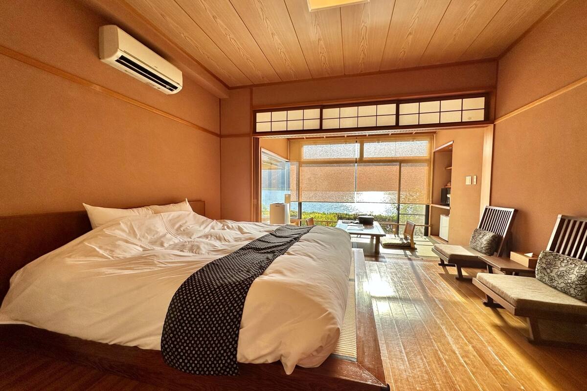 A hotel room with a large bed, a wooden ceiling, and a view through a large window.