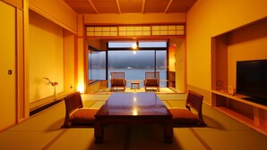 Quarto em estilo japonês tradicional, com tatames, uma mesa baixa, duas cadeiras, uma televisão e vista para a água.