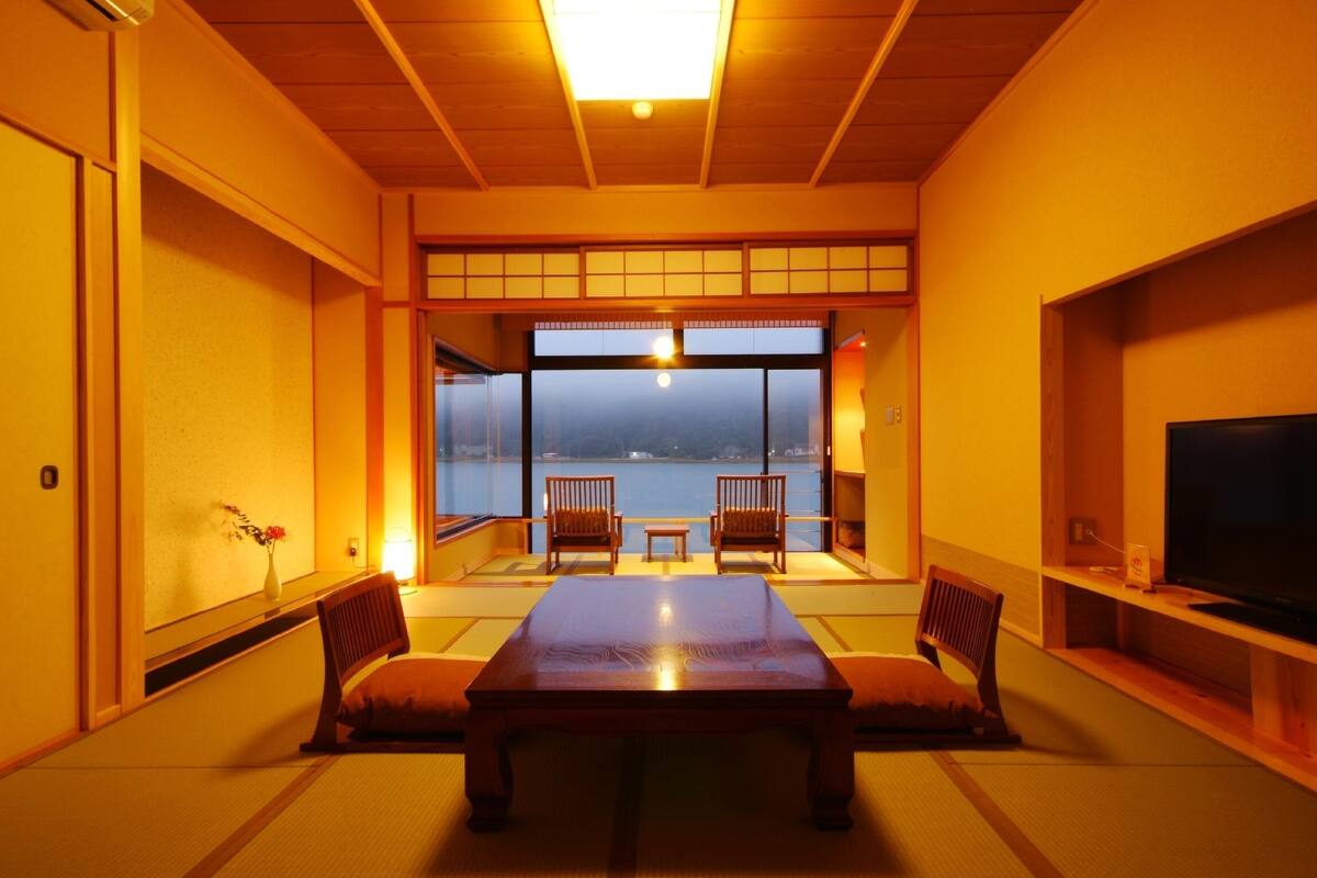 A traditional Japanese-style room with tatami flooring, a low table, two chairs, a television, and a view of a body of water.