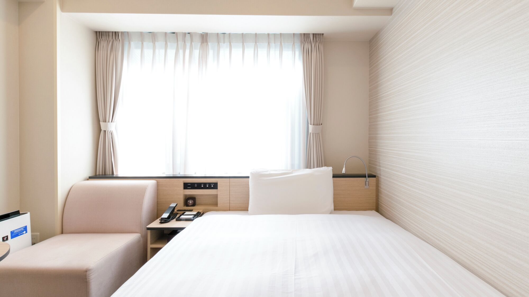 A hotel room with a bed, a desk, a chair, a nightstand, a window with curtains, and a wall with a textured pattern.