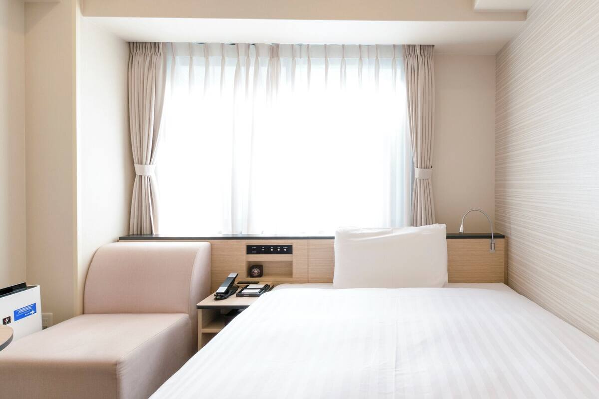 A hotel room with a bed, a desk, a chair, a nightstand, a window with curtains, and a wall with a textured pattern.