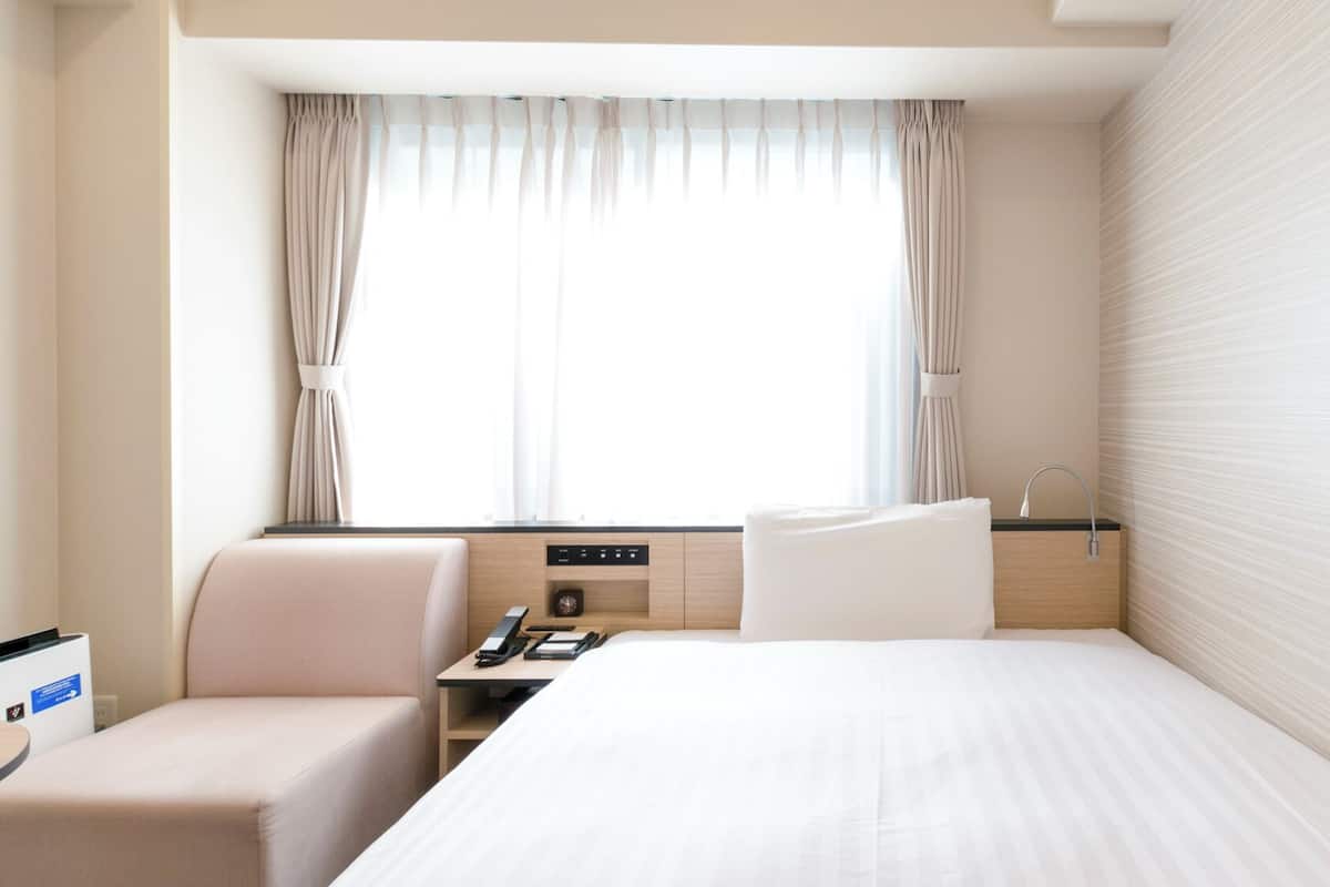 Une chambre d’hôtel comprenant un lit, un bureau, une chaise, une table de chevet, une fenêtre avec des rideaux et un mur orné d’un motif texturé.