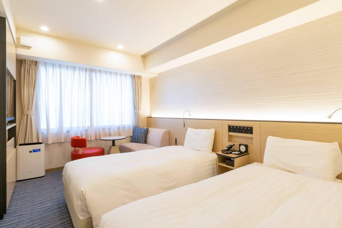 A hotel room with two beds, a small table, a red chair, a desk with a phone and a lamp, and a window with curtains.