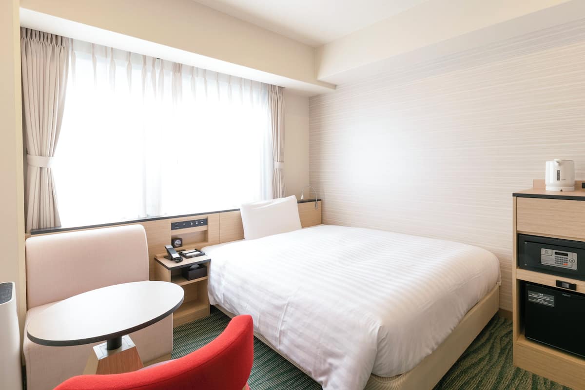 A hotel room with a bed, a desk, a chair, a small table, and a minibar.
