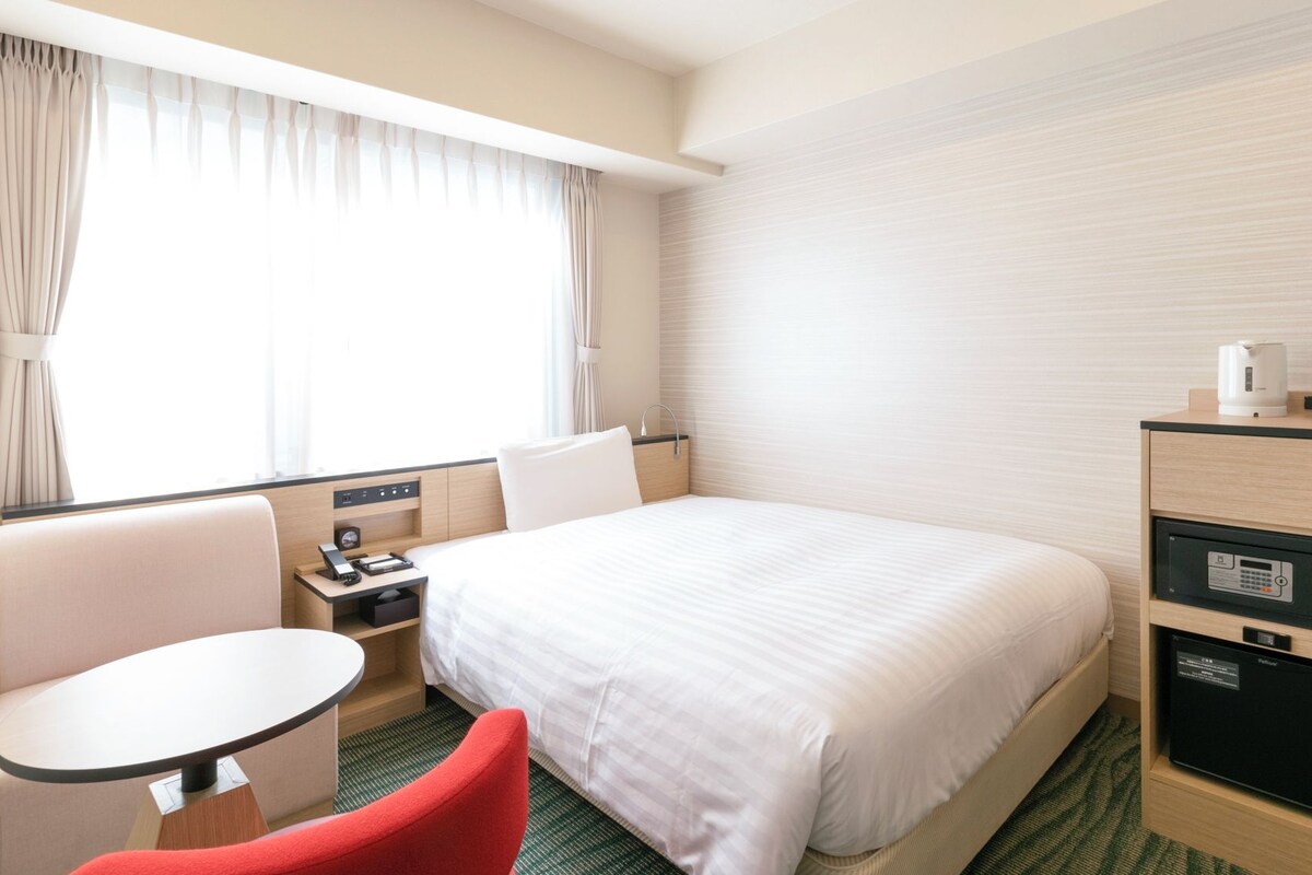 A hotel room with a bed, a desk, a chair, a small table, and a minibar.