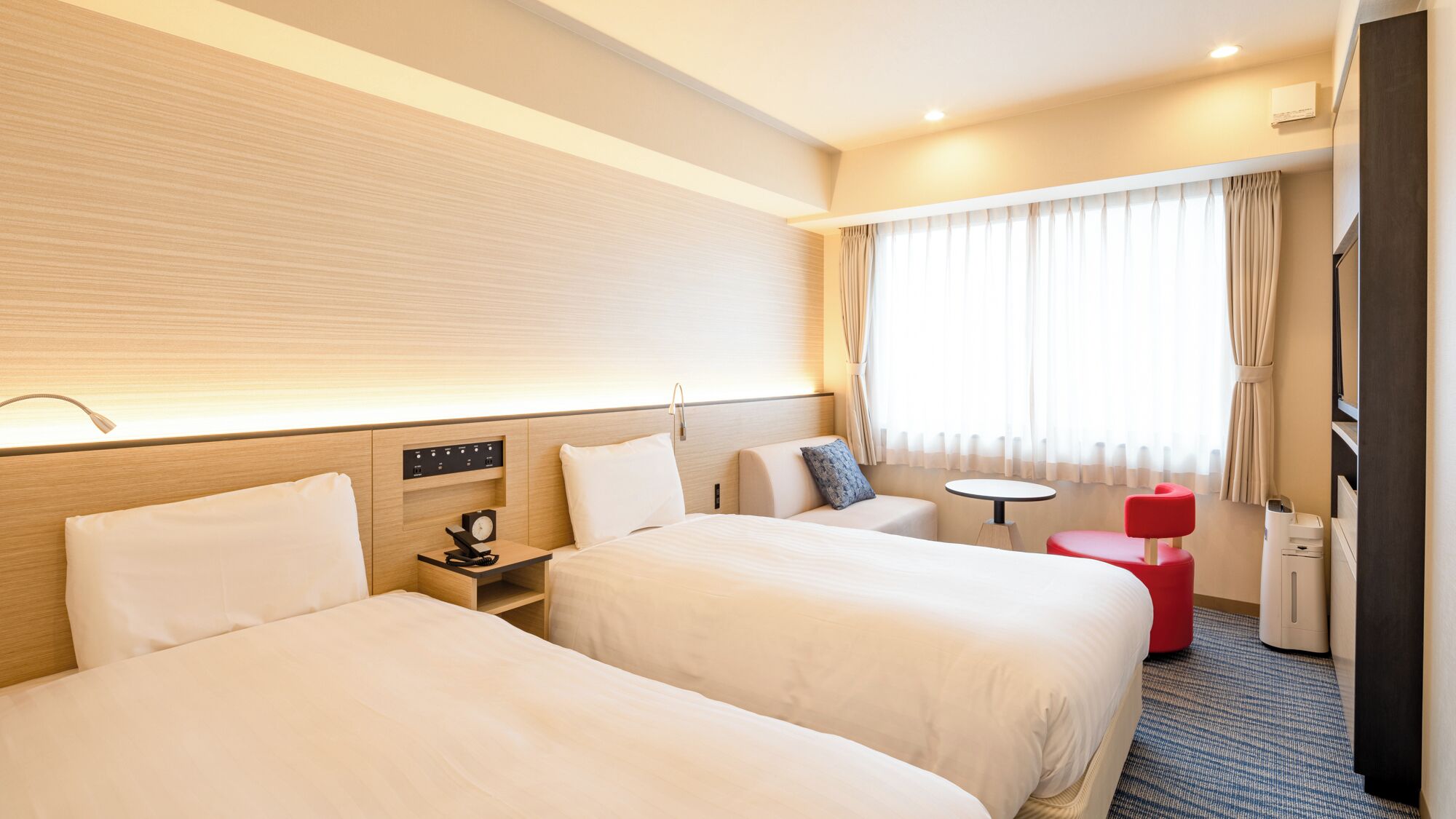 A hotel room with two beds, a small table, a red chair, a desk with a lamp, and a window with curtains.