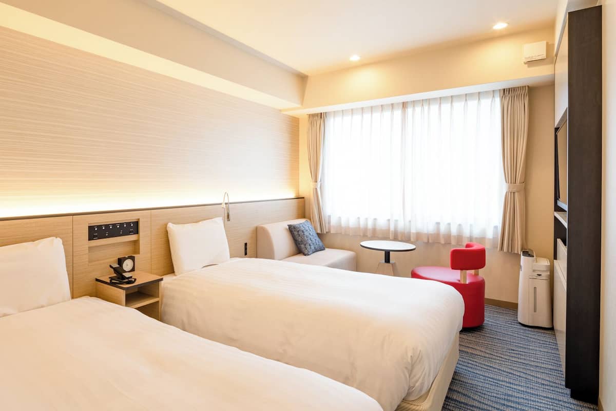 A hotel room with two beds, a small table, a red chair, a desk with a lamp, and a window with curtains.