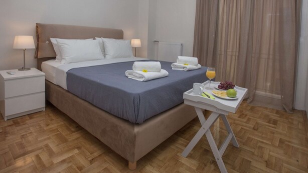 A neatly made bed with white linens and a blue bedspread, a bedside table with a lamp, a tray with fruit and juice, and a window with curtains. - Unique Apartment near Acropolis by GHH (Athens)