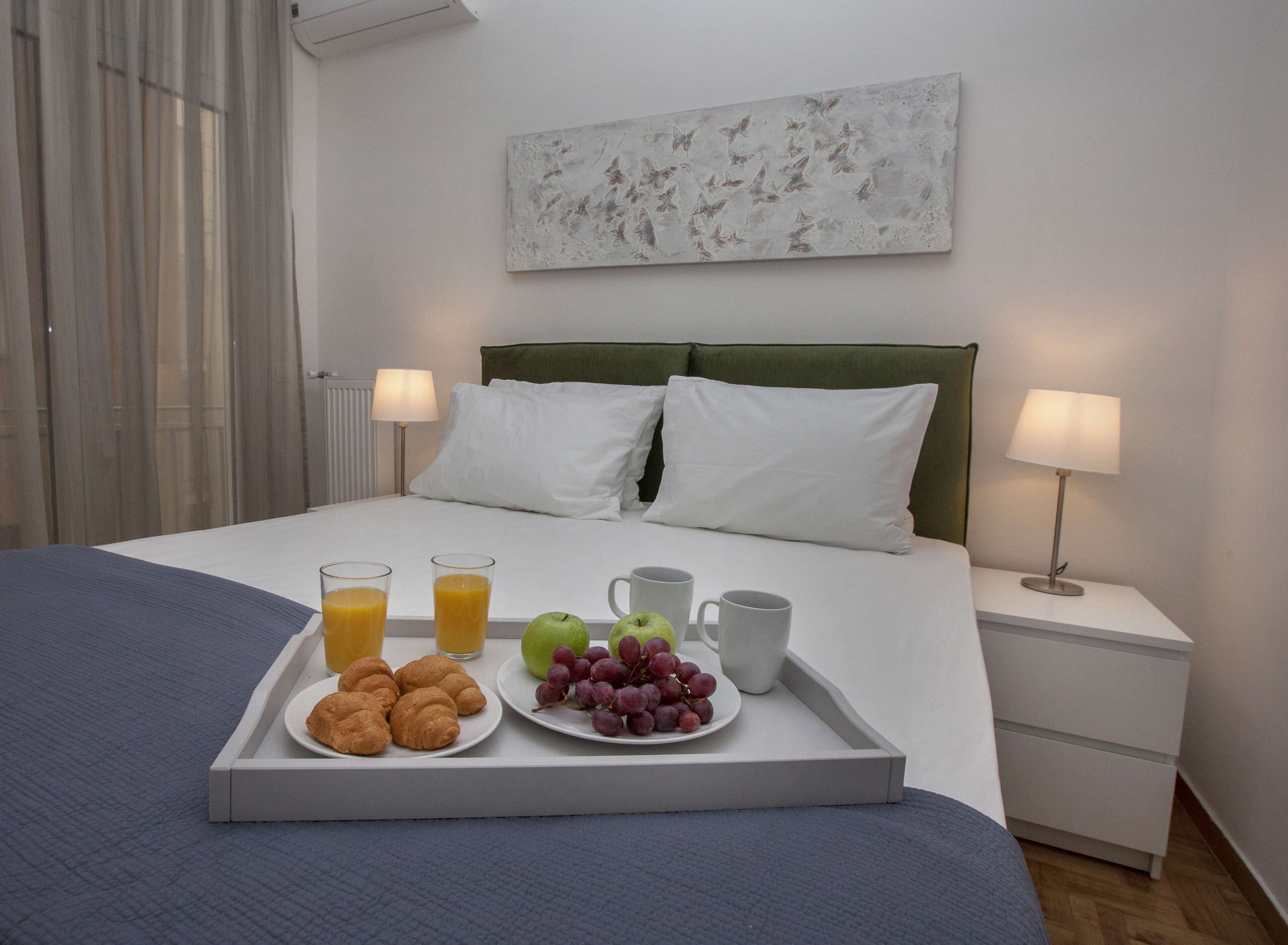 A neatly made bed with white linens, a tray with croissants, fruit, and juice, and two bedside lamps on a nightstand.