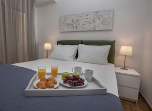 A neatly made bed with white linens, a tray with croissants, fruit, and juice, and two bedside lamps on a nightstand. - Unique Apartment near Acropolis by GHH (Athens)
