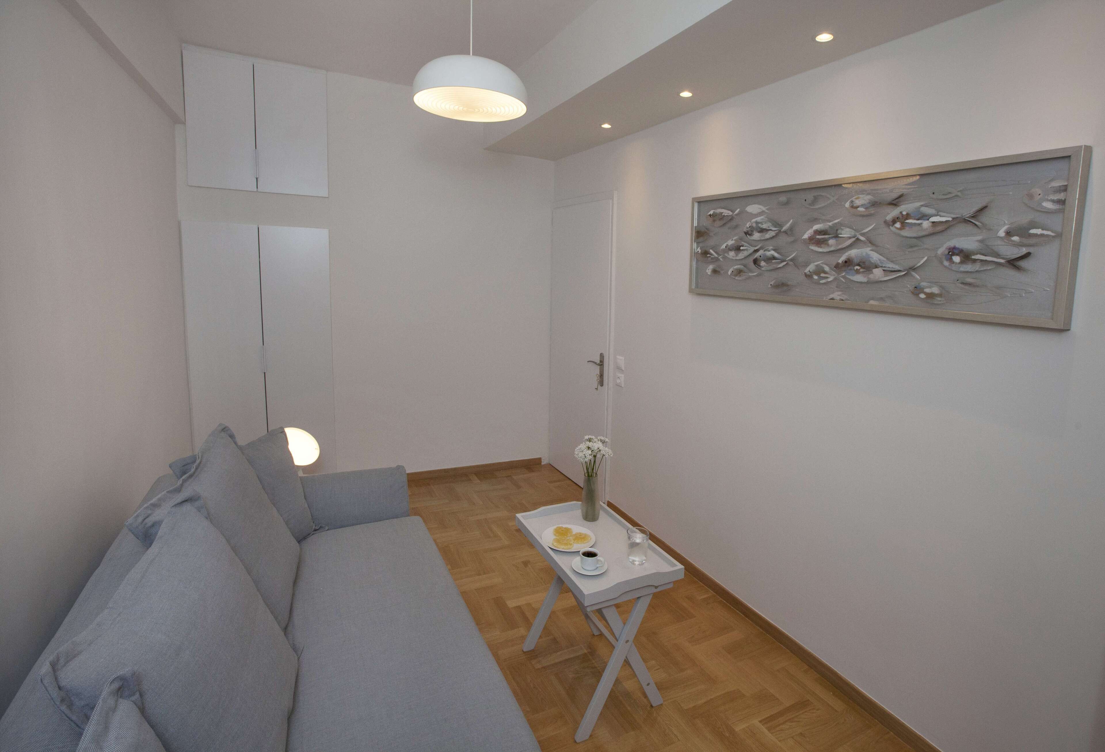 A modern living room with a grey sofa, a small white side table with a tray holding a glass of water and a plate of food, and a wall-mounted fish artwork.
