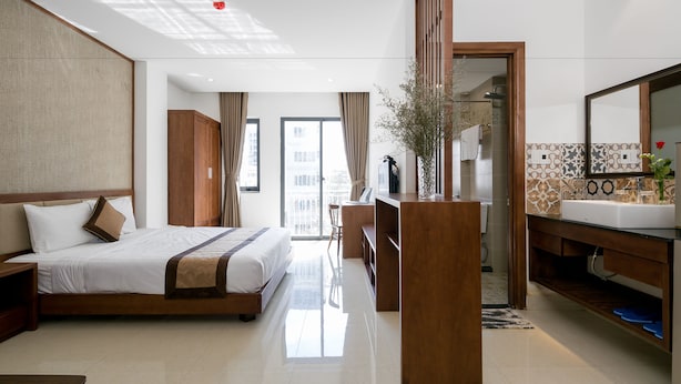 Habitación de hotel con una cama grande, un armario de madera, un baño con espejo y lavamanos, y vistas a la ciudad desde la ventana.