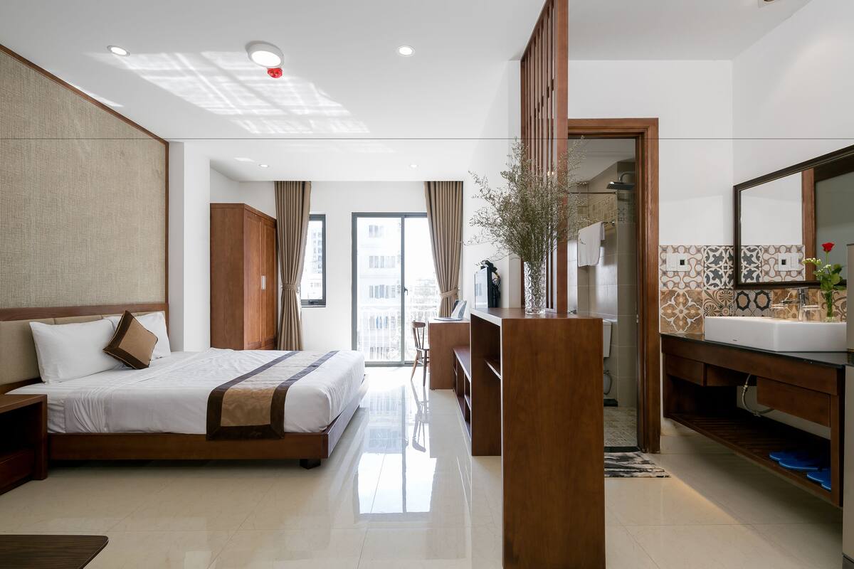 Une chambre d’hôtel avec un grand lit, une armoire en bois, une salle de bain avec un miroir et un lavabo, et une vue sur la ville par la fenêtre.