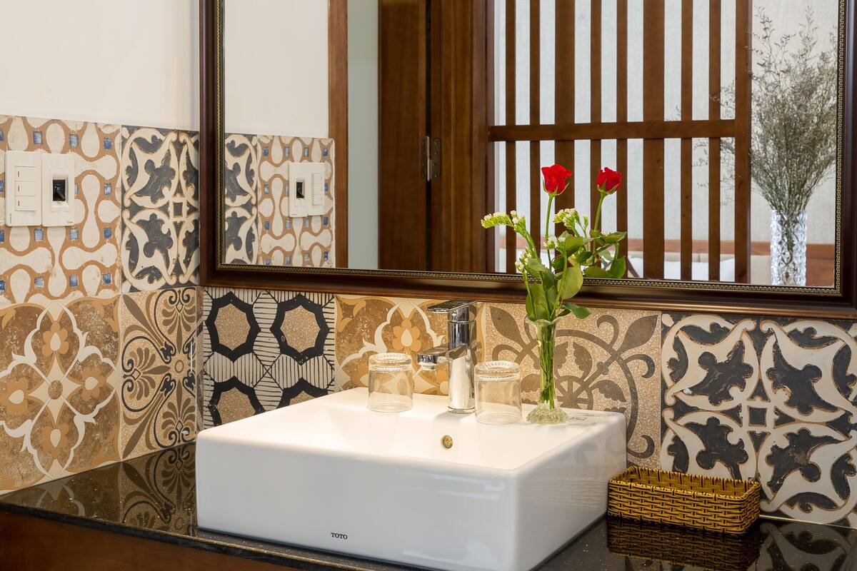 Une salle de bain avec un lavabo blanc, des carreaux décoratifs, un miroir et un vase rempli de fleurs.