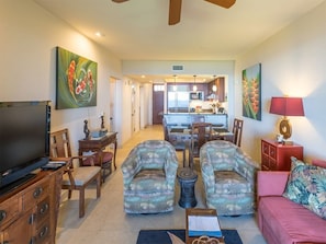 A living room with a TV, two armchairs, a sofa, a dining table, and a kitchen in the background.