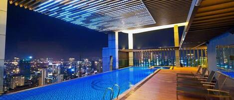 Piscina infinita en la azotea con vista a la ciudad, terraza de madera y sillas de descanso.