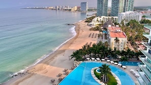 Un resort frente a la playa con piscina, palmeras y edificios altos al fondo.