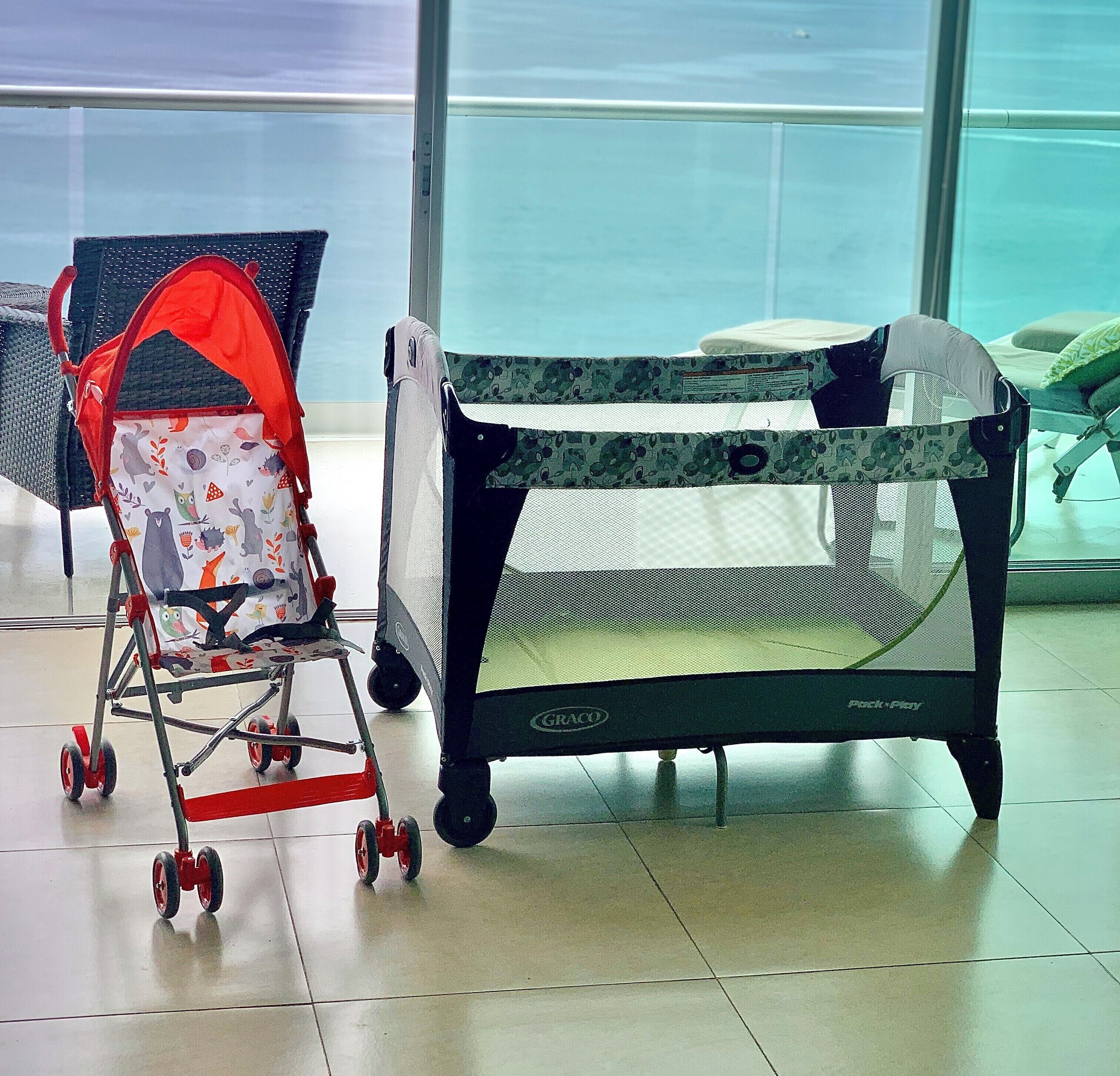 A baby playpen with a mesh top and wheels, a stroller with a red canopy, and a window with a view of the sky.