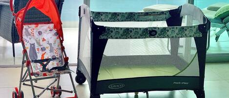 A baby playpen with a mesh top and wheels, a stroller with a red canopy, and a window with a view of the sky.
