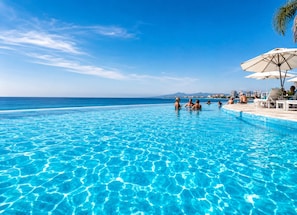 A clear blue pool with people swimming and relaxing, surrounded by lounge chairs and umbrellas, with a view of the ocean and distant buildings.