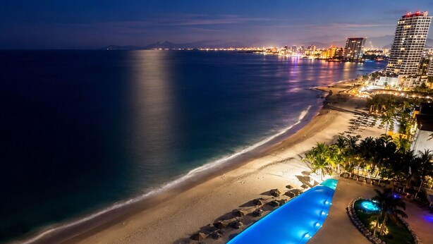 Un complejo turístico en la playa con piscina, palmeras y un horizonte urbano al fondo.