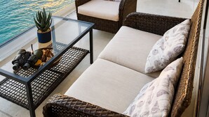 Un balcón con mobiliario de mimbre, una mesa de cristal y vistas a la puesta de sol sobre el océano.