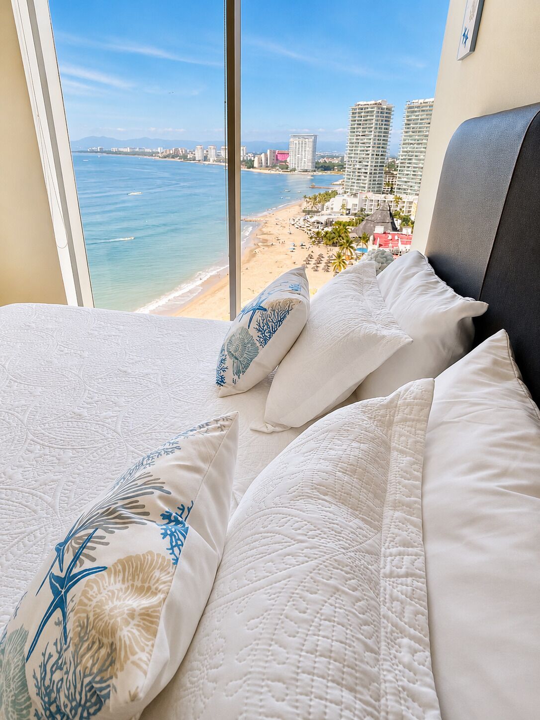 Una cama con ropa de cama blanca y almohadas decorativas, con vistas a una playa y al paisaje urbano a través de una ventana.