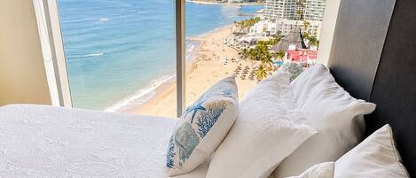 Una cama con ropa de cama blanca y almohadas decorativas, con vistas a una playa y al paisaje urbano a través de una ventana.