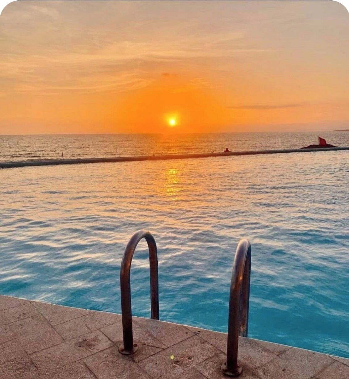 A swimming pool with metal railings, a sunset in the background, and a calm sea.
