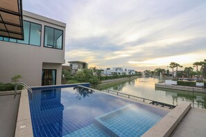 A modern hotel with a rooftop infinity pool overlooking a canal, surrounded by greenery and other buildings.