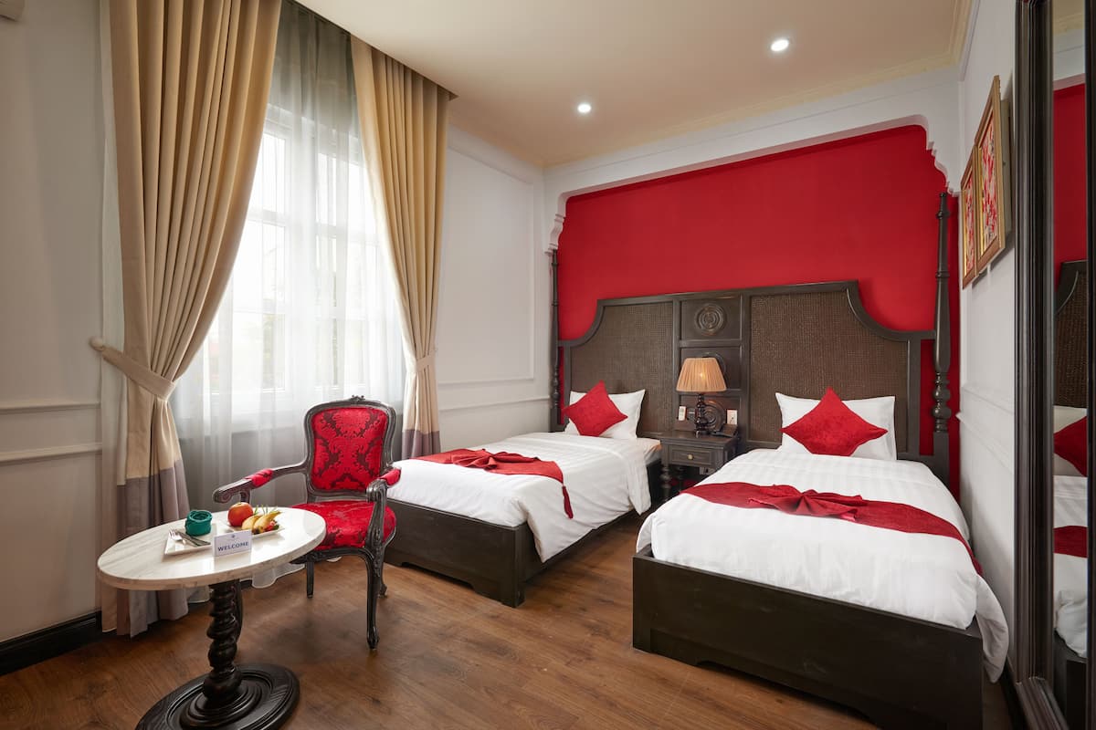 Een hotelkamer met twee bedden, een rode stoel, een klein rond tafeltje met een welkomstbord en een raam met gordijnen.