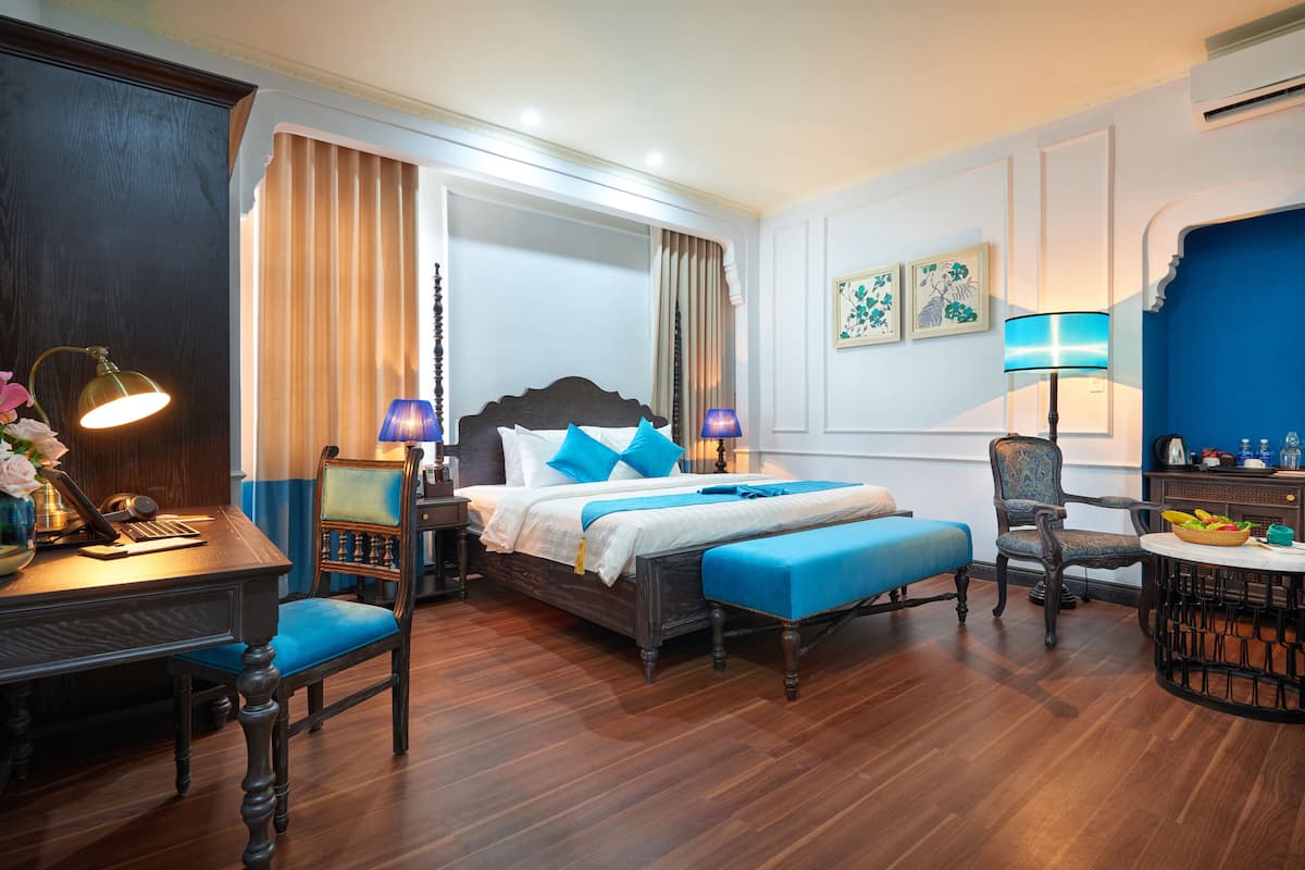 Een hotelkamer met een houten bed, een blauw gestoffeerde stoel, een bureau met een lamp en een ronde tafel met een schaal fruit.